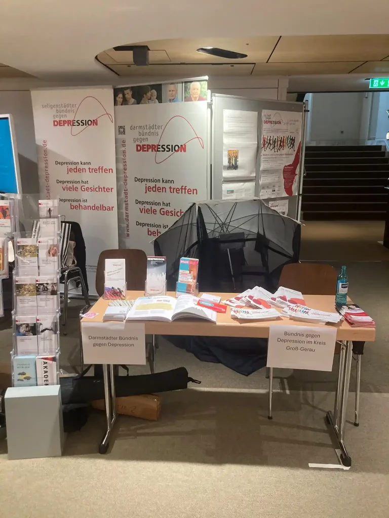 Ein gut ausgestatteter Informationsstand mit Flyern, Broschüren und großen Bannern der Bündnisse gegen Depression aus Darmstadt, Seligenstadt und dem Kreis Groß-Gerau in einem Veranstaltungsfoyer.