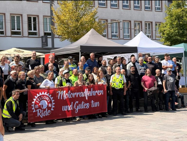 Eine große Gruppe von Menschen posiert gemeinsam auf dem Darmstädter Friedensplatz hinter einem roten Banner mit der Aufschrift "Fellows Ride - Motorradfahren und Gutes tun".